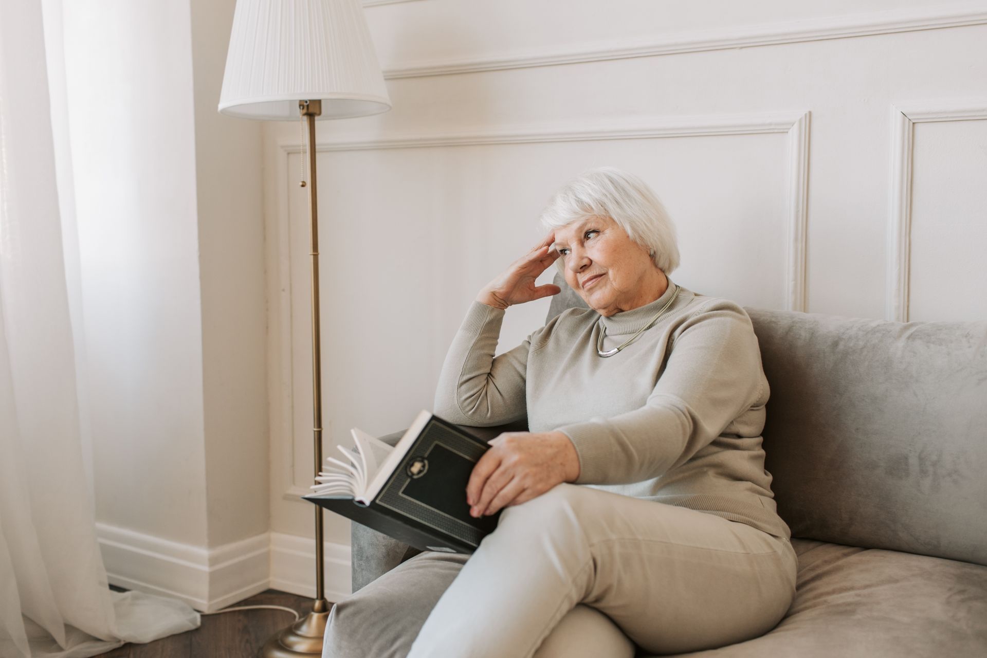 Senior woman with grey hair sitting on a couch reading a book, thoughtful expression