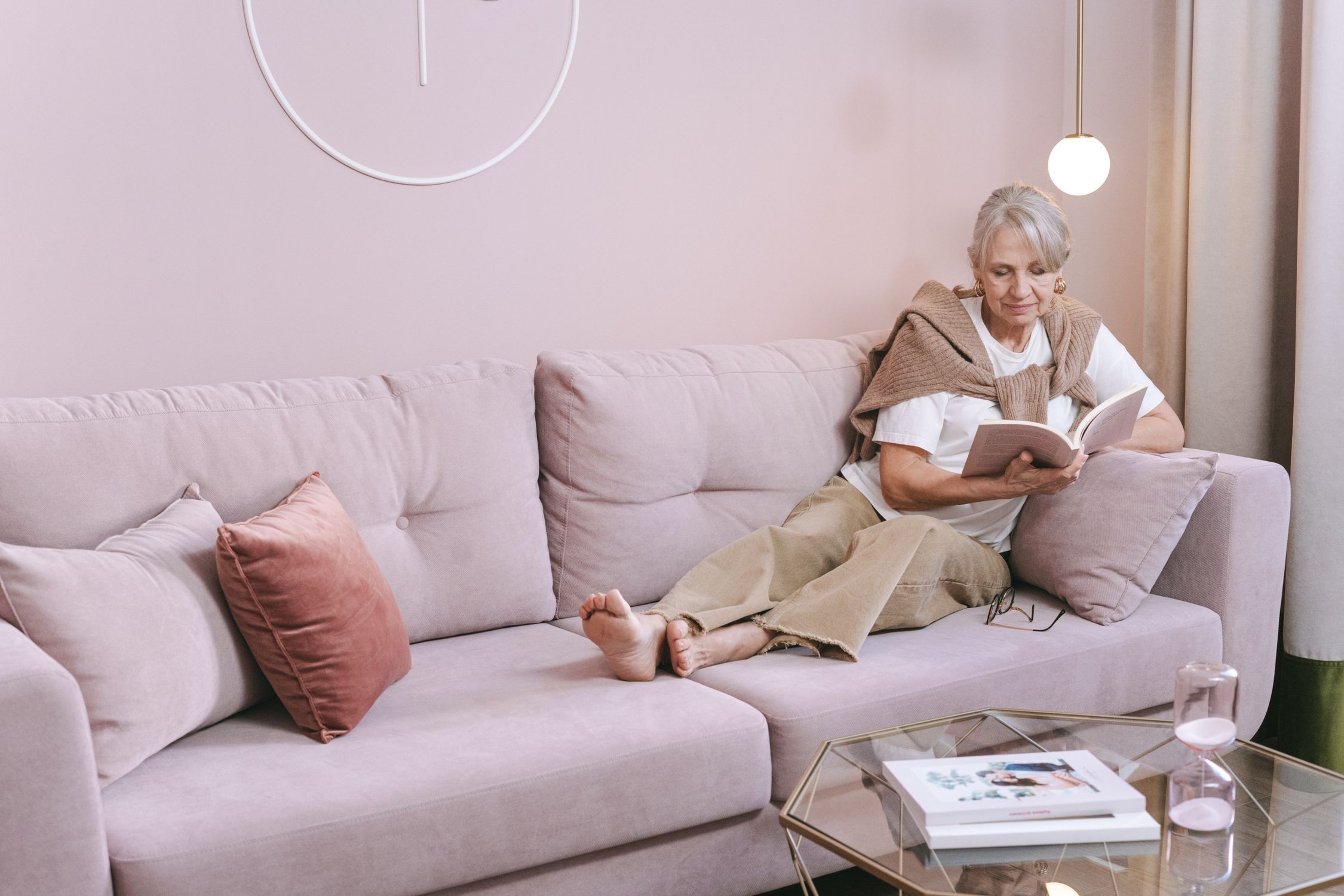 Senior woman with grey hair reading a book on a comfortable pink sofa at home