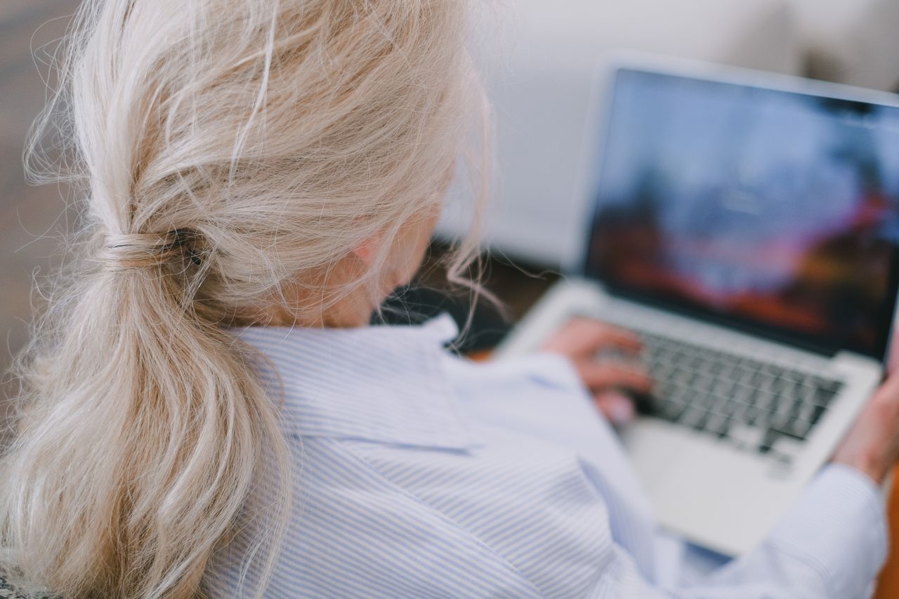 Older woman with white hair typing on a laptop, seen from behind in a striped shirt