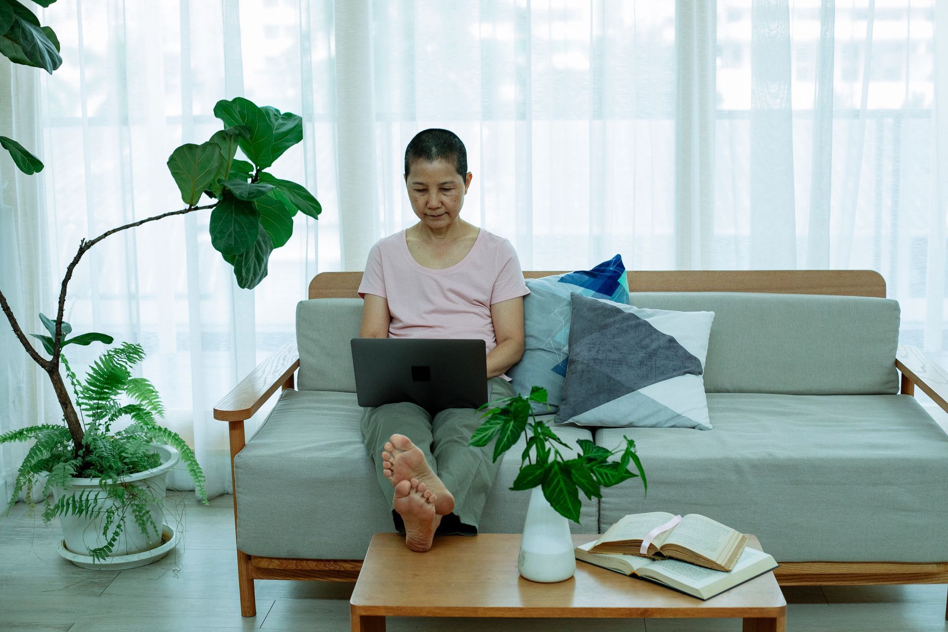 Mature Asian woman sitting on a couch with pillows browsing on a laptop at home