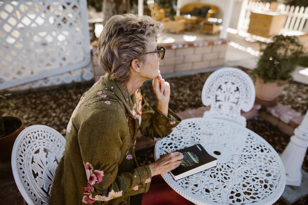 Older woman with glasses sitting at a garden table with a book, looking away in thought