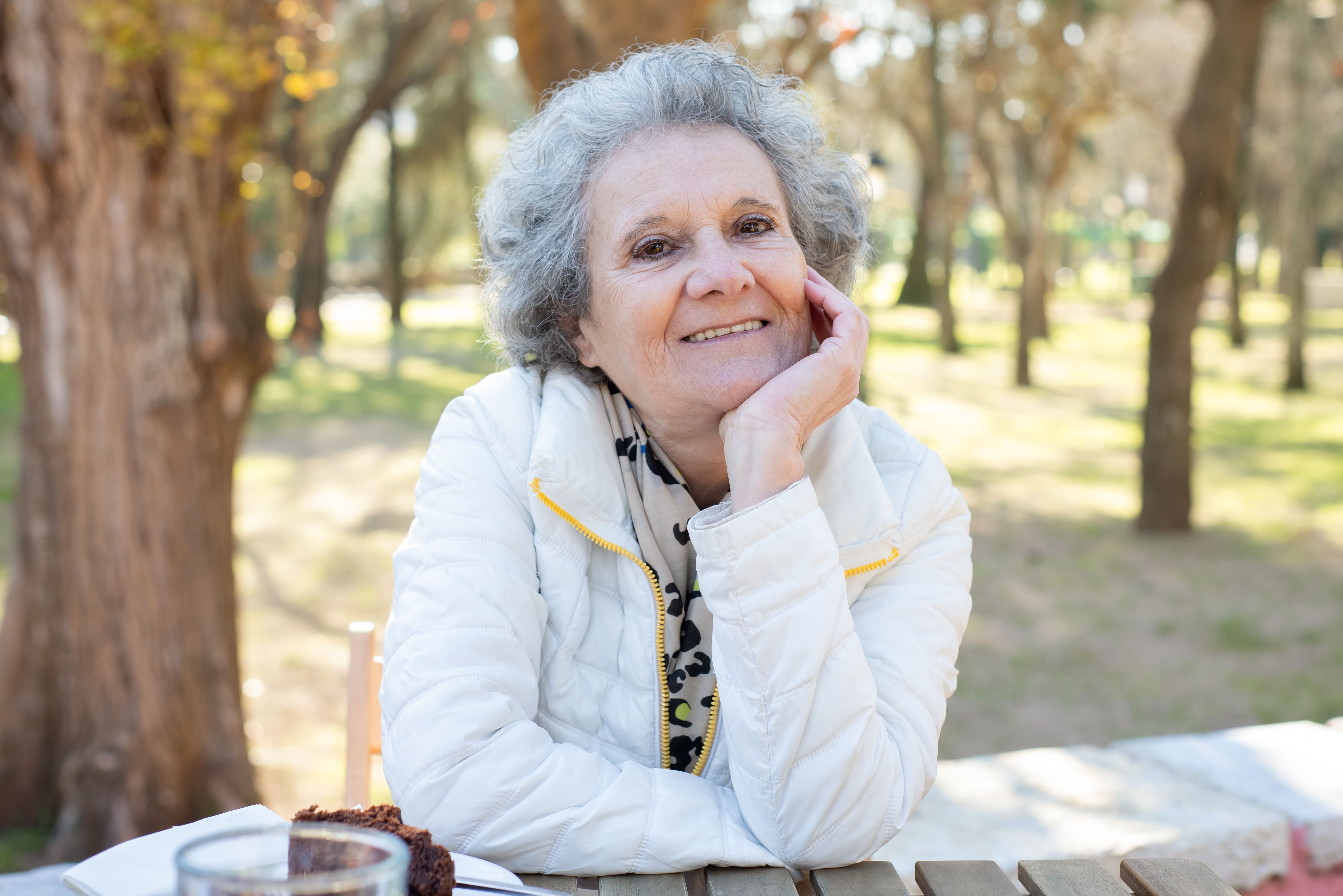 Woman in her sixties with gray hair sitting at a park table, hand resting on cheek, warm afternoon light