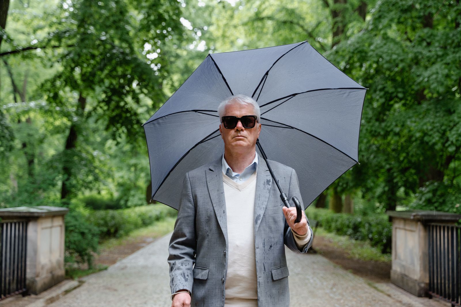 Silver-haired man in a gray blazer holding an umbrella on a tree-lined park path