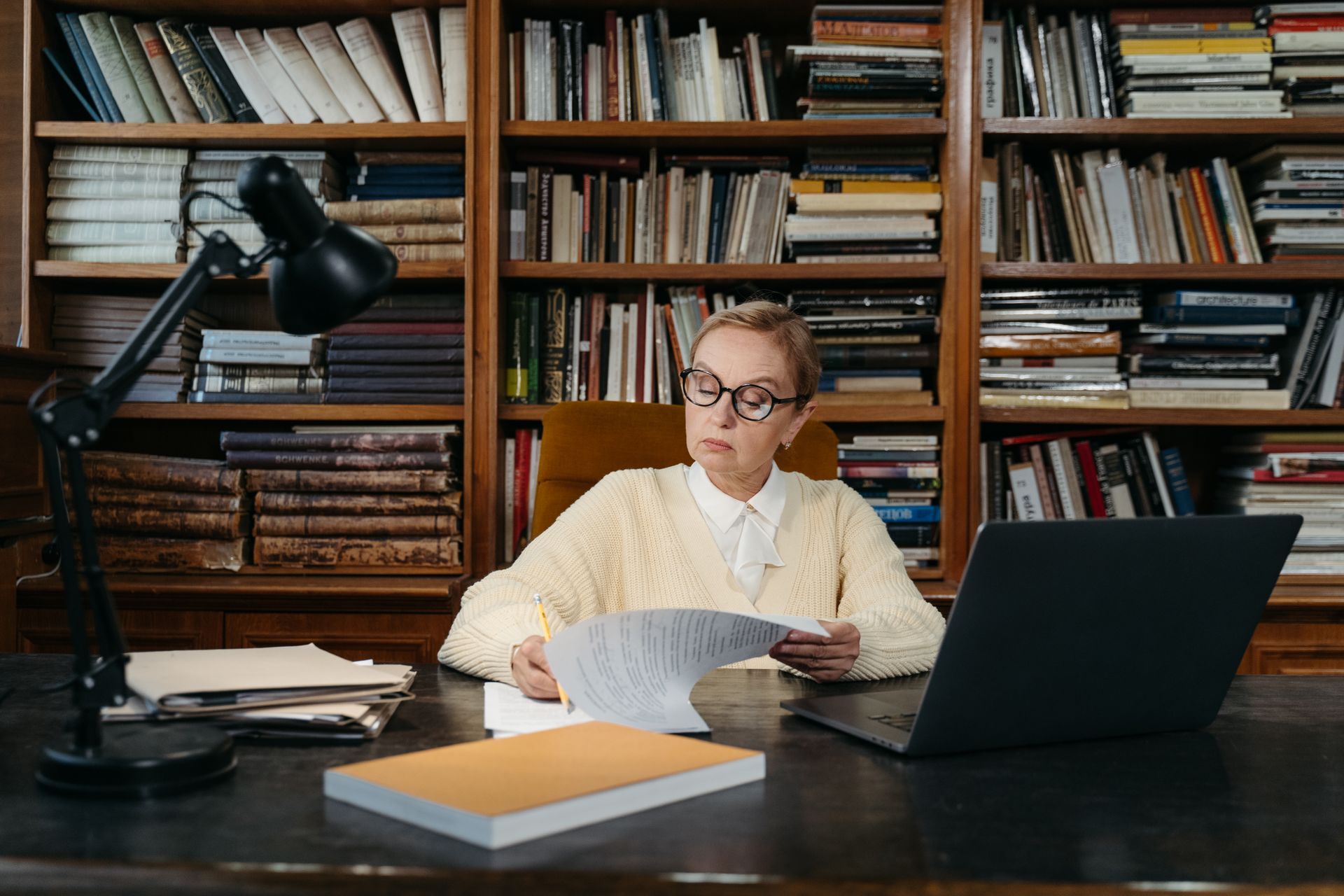 Senior woman with grey hair reading documents at a library desk with books
