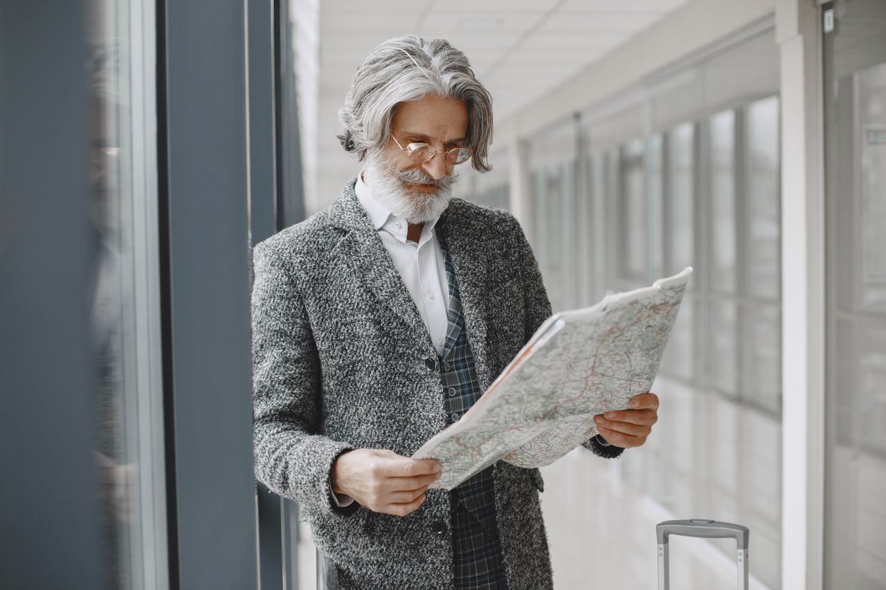 Gray-haired man with glasses reading a map in an airport corridor with a suitcase beside him