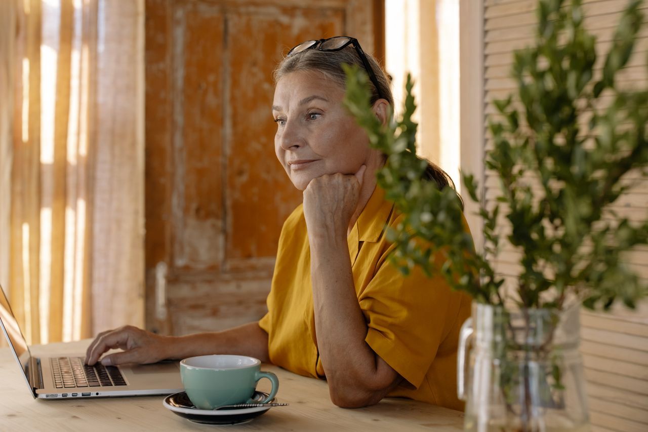 Older woman resting her chin on one hand at a wooden table with a laptop and cup in warm afternoon light