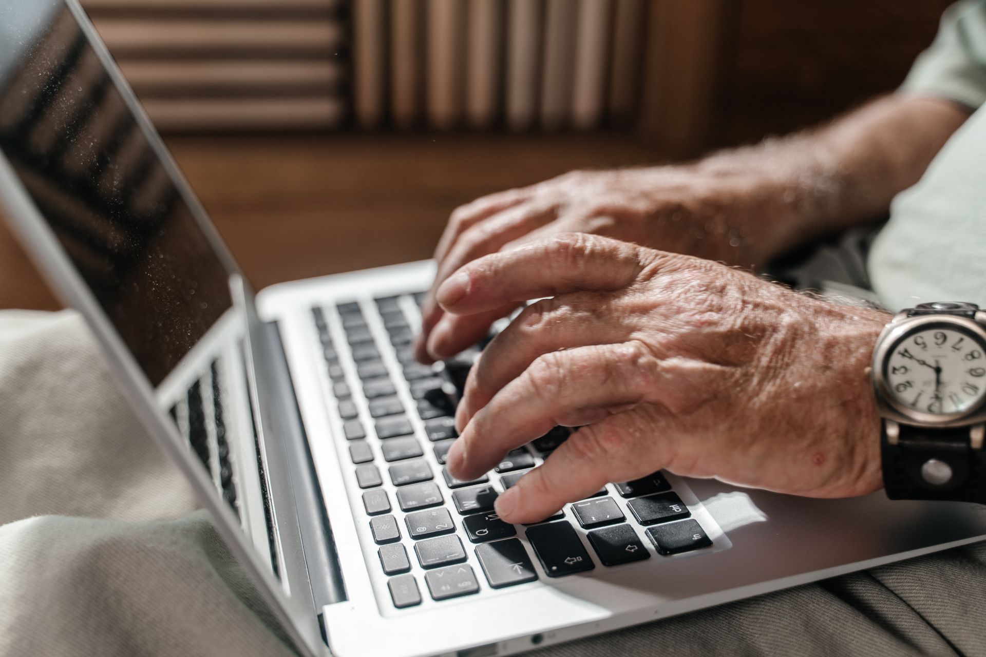 Older hands typing on a laptop keyboard at home in warm natural light