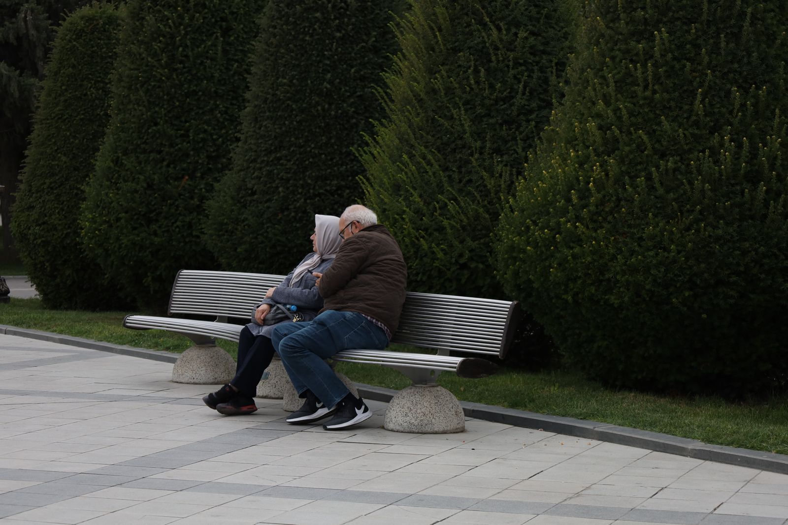 Older couple sitting together on a park bench surrounded by green hedges, looking in the same direction