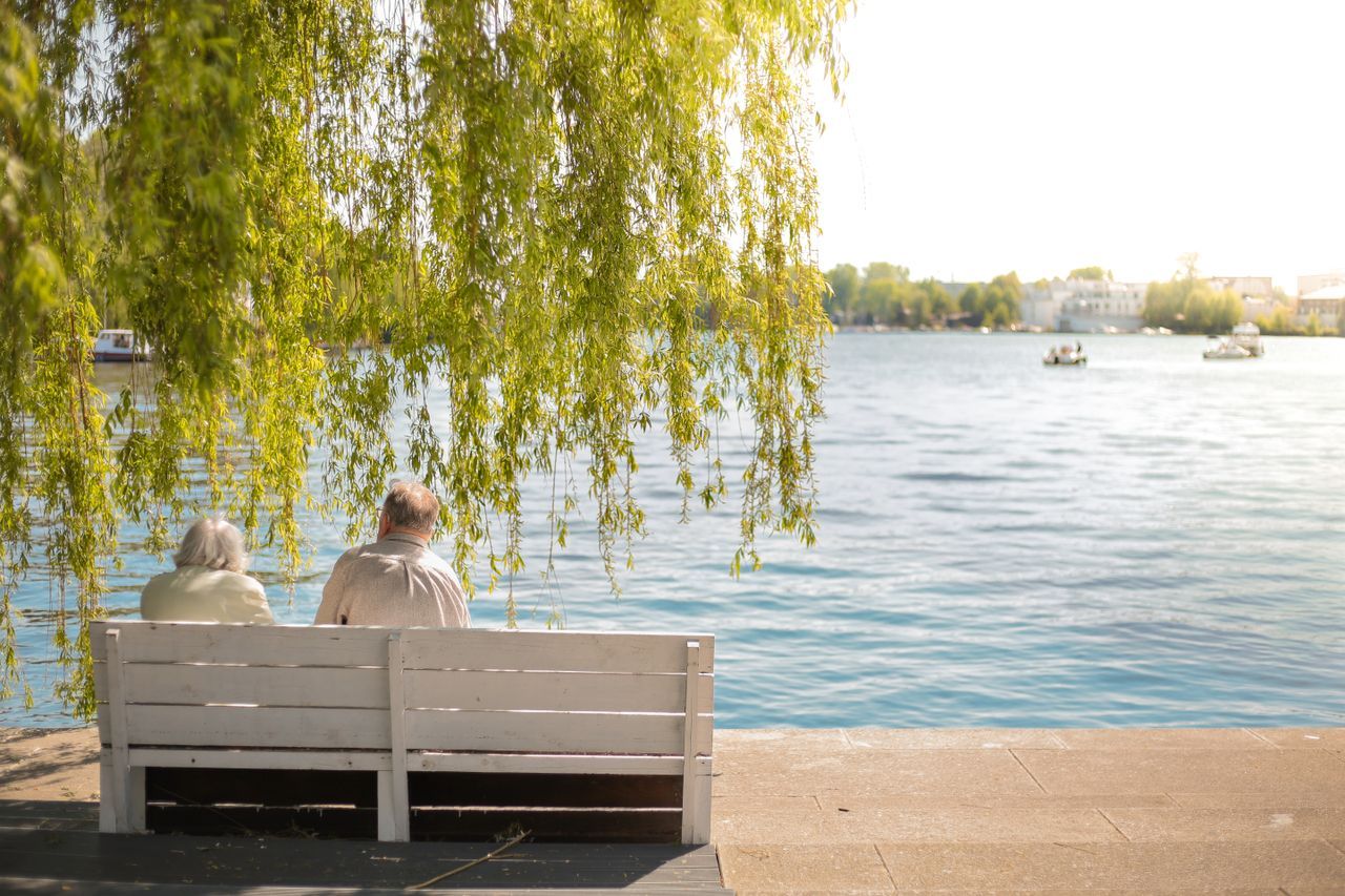 Elderly couple sitting together on a bench by a lake under a willow tree