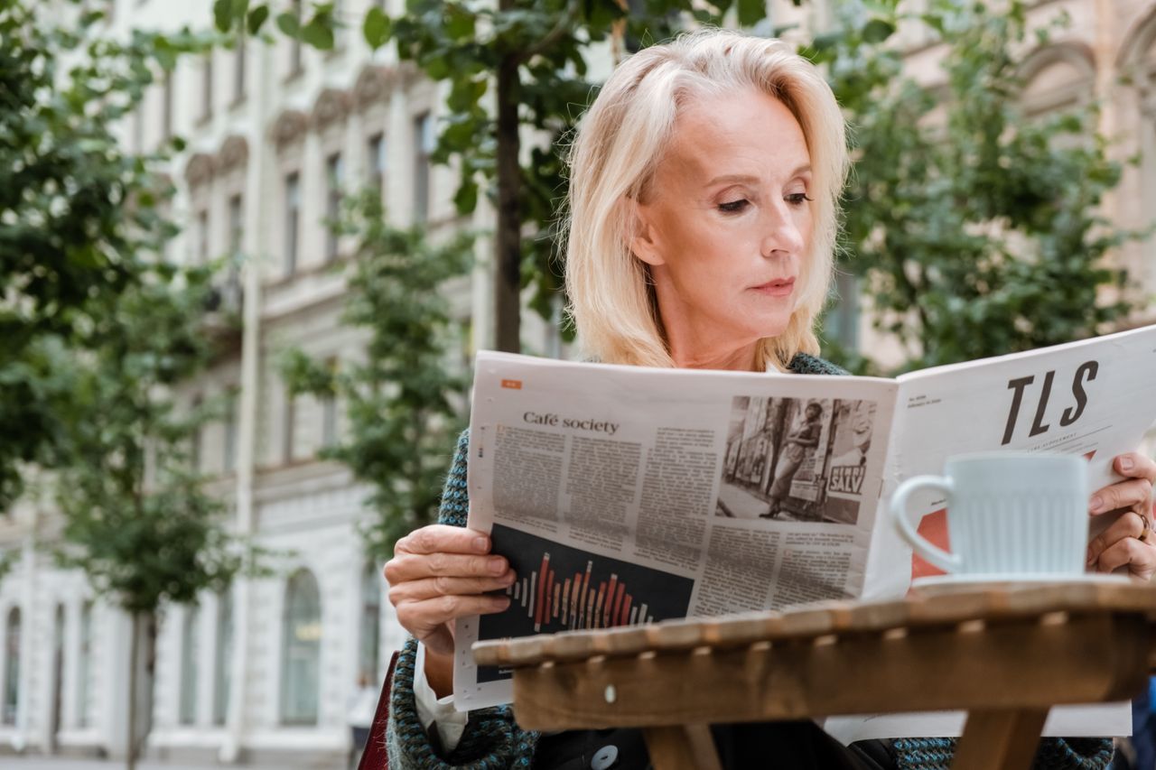 Mature woman reading a newspaper alone at an outdoor café table on a bright summer morning