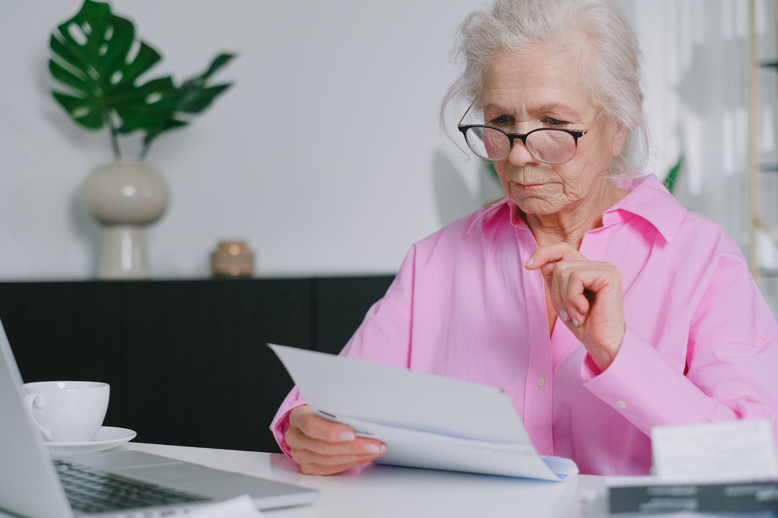 Elderly woman in pink shirt reviewing documents at a home desk with laptop and coffee