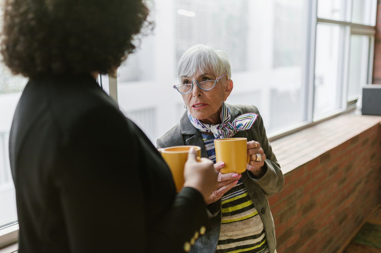 Older woman with white hair and glasses holding a coffee mug in calm conversation indoors