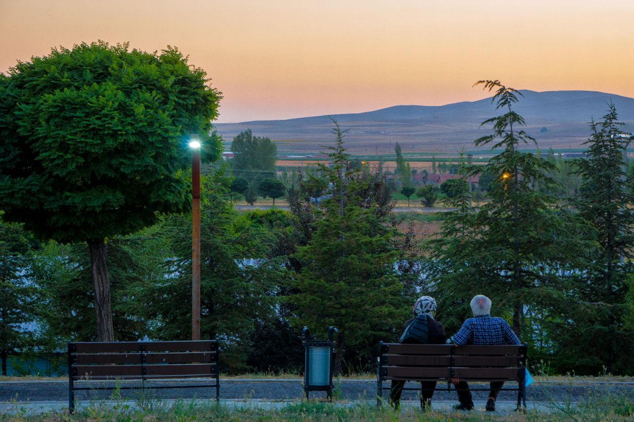 Elderly couple sitting together on a park bench at sunset, seen from behind, looking out over an open landscape with mountains