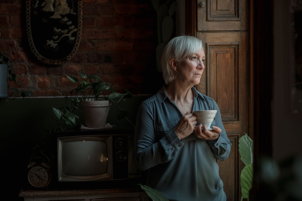 Senior woman with grey hair gazing out a window holding a cup, surrounded by houseplants