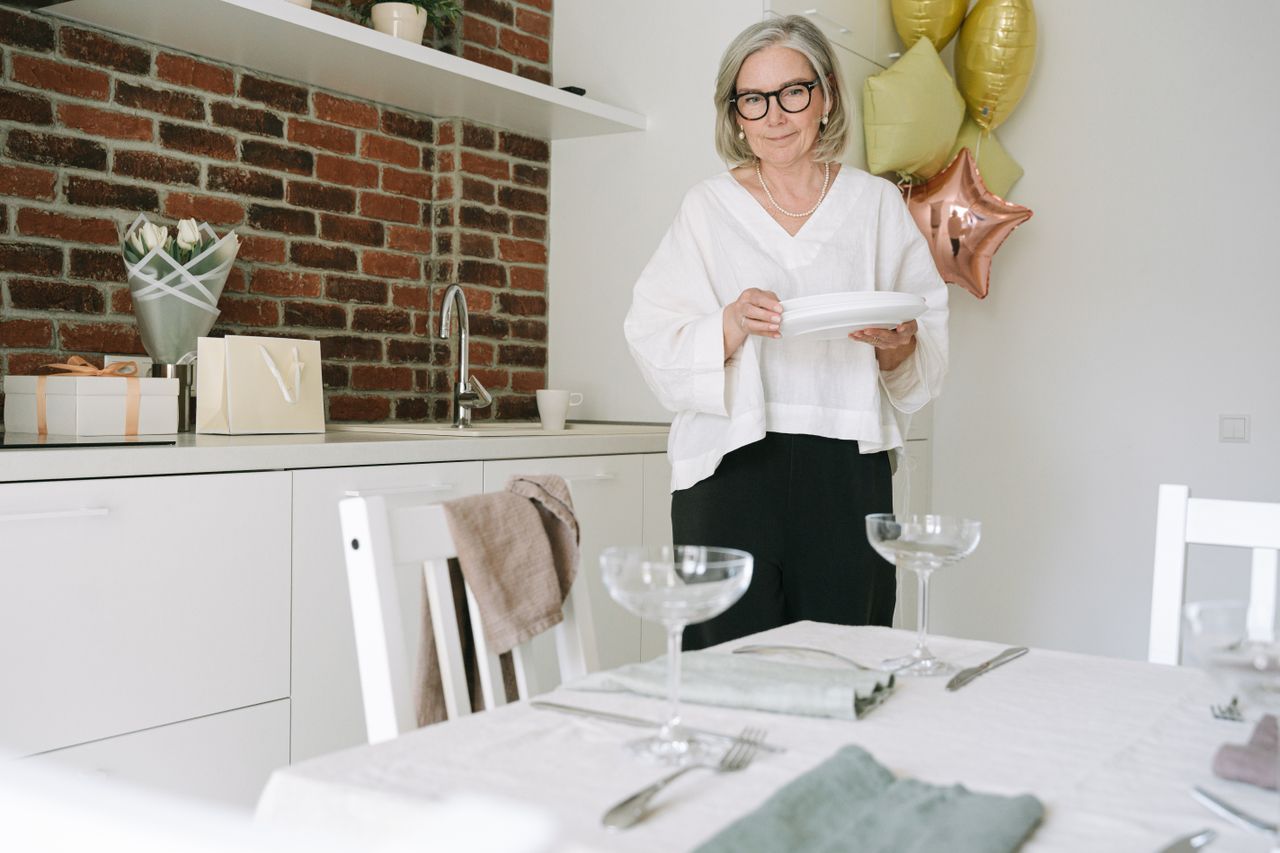 Woman over 50 with gray hair setting plates on a table prepared for a small gathering in a modern kitchen