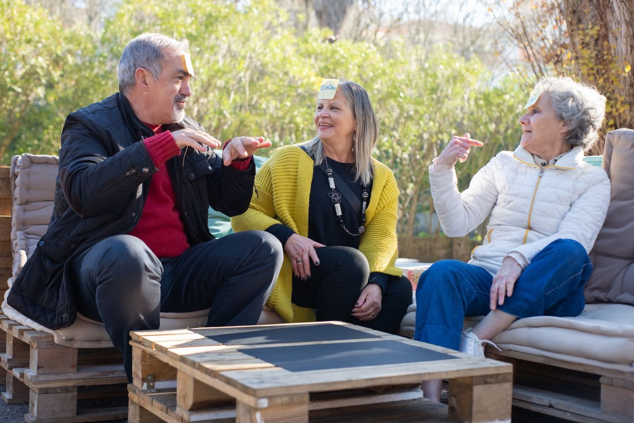 Three older adults laughing together while playing a guessing game outdoors in a garden