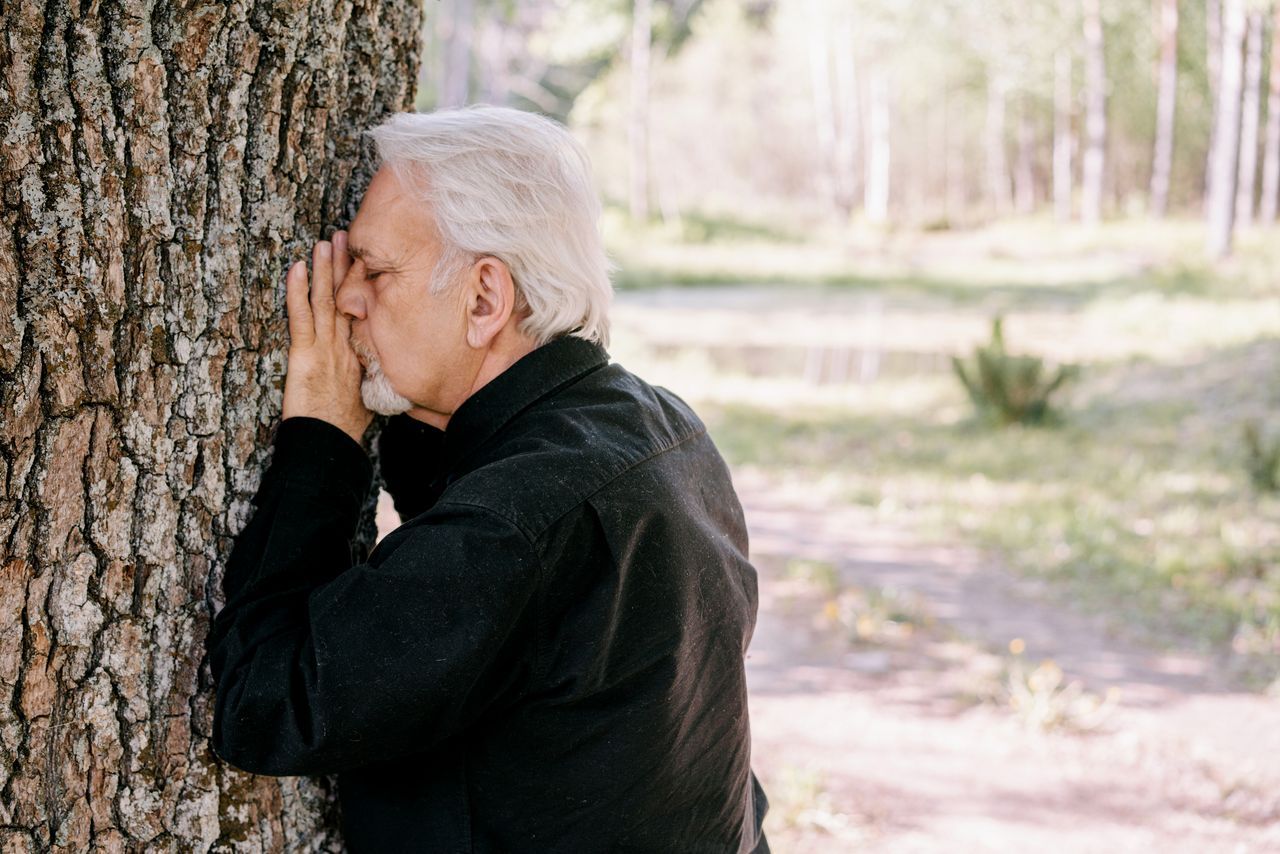Senior man in a dark shirt leaning against a tree trunk outdoors, looking thoughtful